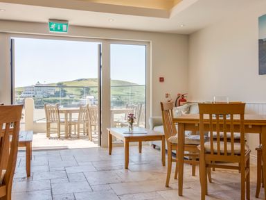 A dining area with wooden tables and chairs next to a glass door opening to an outdoor seating area overlooking hills at 4 Burgh Island Causeway Bigbury