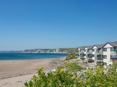 A beach with a few people walking along the shore next to a row of houses with glass balconies at 4 Burgh Island Causeway Bigbury