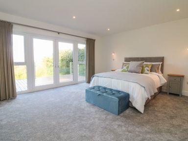 A bedroom with a bed, two bedside tables, a blue storage bench, and large windows with curtains at Linden View in Combe Martin