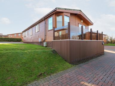 A wooden cabin with a glass railing on the deck and green grass beside a brick pathway at Serenity Sunset in Ilfracombe