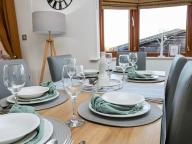 A dining table set with plates glasses and napkins in a room with a floor lamp and window at Serenity Sunset in Ilfracombe