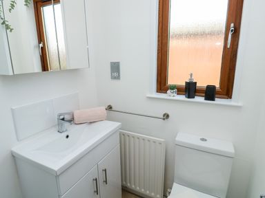 A bathroom with a white sink cabinet and a toilet under a window with accessories on the sill at Serenity Sunset in Ilfracombe