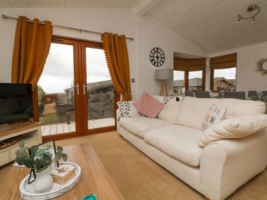 A living room with a cream sofa pink cushions a wooden coffee table and glass doors with orange curtains at Serenity Sunset in Ilfracombe