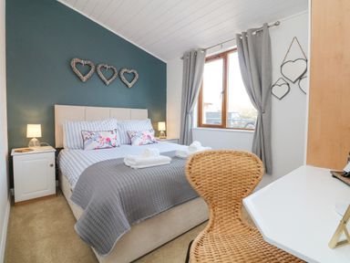 A bedroom with a double bed, bedside tables with lamps, heart decorations on walls, a window with gray curtains, and a wicker chair at Serenity Sunset in Ilfracombe