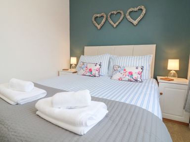 A bedroom with a bed with striped bedding and floral pillows two bedside tables with lamps and woven heart decorations on the wall at Serenity Sunset in Ilfracombe