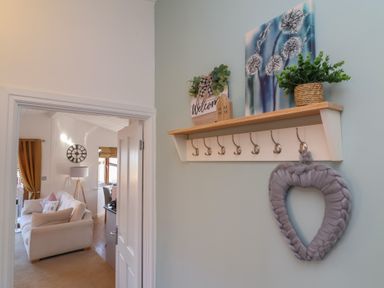A hallway wall with a shelf and hooks with decor and a heart-shaped wreath and a view of a living room with a sofa and floor lamp at Serenity Sunset in Ilfracombe