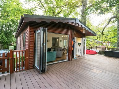 A wooden cabin exterior with an open patio door showing a living area and a wooden deck with outdoor items at Tranquil Waters in Lakeside 16 White Cross Bay near Windermere