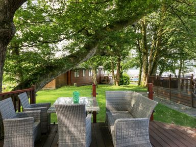 An outdoor seating area with wicker chairs and a glass table on a wooden deck surrounded by trees and a wooden cabin at Tranquil Waters in Lakeside 16 White Cross Bay near Windermere