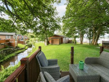 An outdoor seating area with chairs and a table overlooking a grassy area with trees and wooden cabins by a small stream at Tranquil Waters in Lakeside 16 White Cross Bay near Windermere