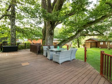 An outdoor deck with a glass table and wicker chairs surrounded by large trees and wooden cabins at Tranquil Waters in Lakeside 16 White Cross Bay near Windermere