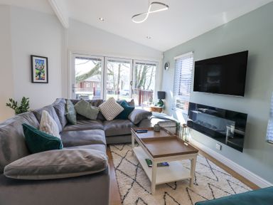 A living room with a gray sectional sofa green and white cushions a wooden coffee table a wall-mounted tv and glass doors at Tranquil Waters Lakeside 16 White Cross Bay near Windermere