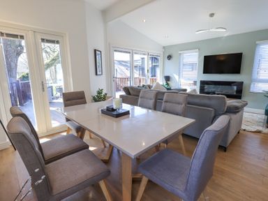 A dining area with a table and six chairs next to a living room with sofas and a wall-mounted TV at Tranquil Waters Lakeside 16 White Cross Bay near Windermere