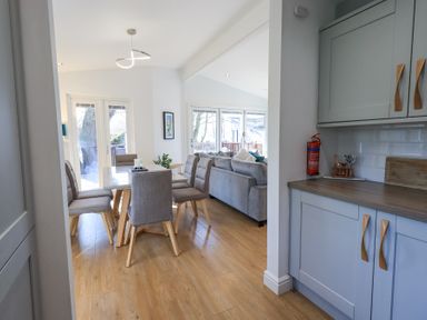 A dining area with gray chairs and a table adjacent to a seating area with a sofa and kitchen cabinets at Tranquil Waters Lakeside 16 White Cross Bay near Windermere