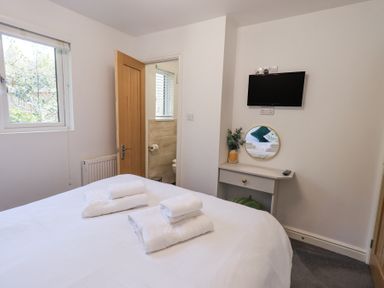 A bedroom with a bed, white towels on the bed, a small table with a mirror and a vase, a wall-mounted TV, and an open door showing a bathroom at Tranquil Waters Lakeside 16 White Cross Bay near Windermere