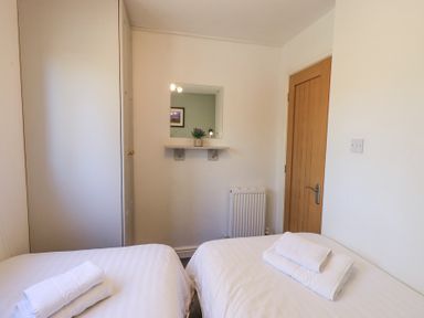 A bedroom with two single beds with white linens and towels a mirror above a shelf on the wall and a wooden door at Tranquil Waters in Lakeside 16 White Cross Bay near Windermere