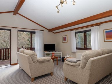A living room with two beige sofas a TV on a stand a coffee table and windows with white curtains at Teign River Retreat in Drewsteignton near Moretonhampstead
