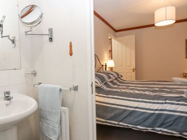 A bedroom with a bed and a bedside lamp visible through an open door from a small bathroom with a sink and towel
