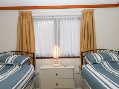 A bedroom with two single beds with striped bedding a white nightstand with a lamp and a book between them and mustard curtains at Teign River Retreat in Drewsteignton near Moretonhampstead
