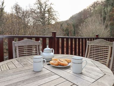 A wooden outdoor table with two white mugs a teapot and a plate of pastries on a balcony with wooden chairs and trees in the background at Teign River Retreat in Drewsteignton near Moretonhampstead