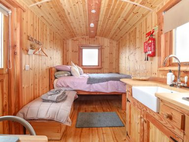 A small wooden room with a bed, bench with cushions, kitchen sink, and window at Hafan Gelli Sheperds Hut - Enlli in Pwllheli