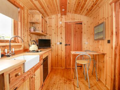 A kitchen with wooden cabinets a sink a kettle small dining table with two chairs and a wall-mounted picture at Hafan Gelli Sheperds Hut - Enlli in Pwllheli