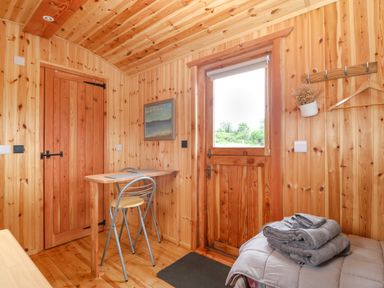 A wooden room with a door a small table with a chair a bed with folded towels a wall hook with a hanger and a potted plant at Hafan Gelli Sheperds Hut - Enlli in Pwllheli
