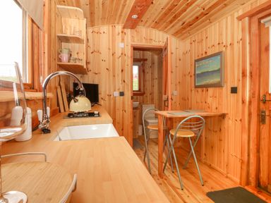 A small wooden kitchen with a sink, stove, dining table with two chairs and a bathroom visible through a door at Hafan Gelli Sheperds Hut - Enlli in Pwllheli
