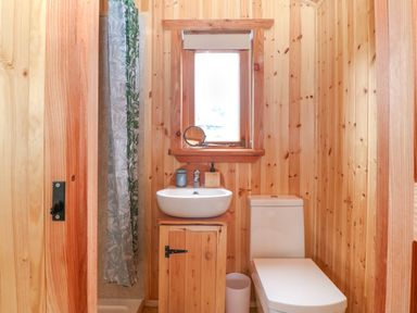 A small bathroom with wooden walls a white sink a toilet a window and a shower curtain at Hafan Gelli Sheperds Hut - Enlli in Pwllheli