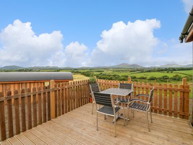 A wooden deck with a metal table and four chairs surrounded by a wooden fence and a grassy landscape at Hafan Gelli Sheperds Hut - Enlli in Pwllheli