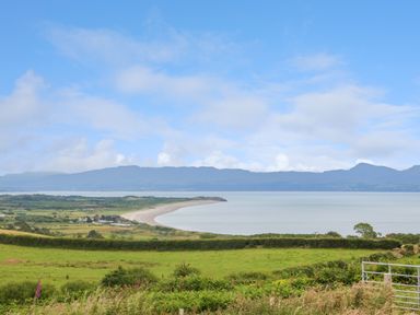 A coastal landscape with green fields and bushes in the foreground and a bay with mountains in the background at Hafan Gelli Sheperds Hut - Enlli in Pwllheli