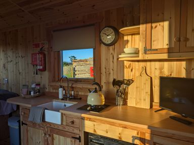 A kitchen area with a sink kettle utensils clock and small tv at Hafan Gelli Sheperds Hut - Enlli in Pwllheli