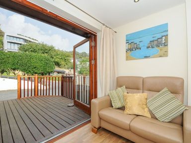 A seating area with a beige leather sofa with cushions and an open glass door leading to a wooden deck outside at Little Trevallan in Looe