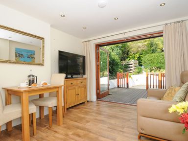 A living room with a wooden dining table and chairs a flat screen tv on a cabinet a sofa and open doors leading to a garden at Little Trevallan in Looe