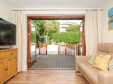 A living room with a leather sofa and wooden cabinet next to open double doors leading to a deck at Little Trevallan in Looe