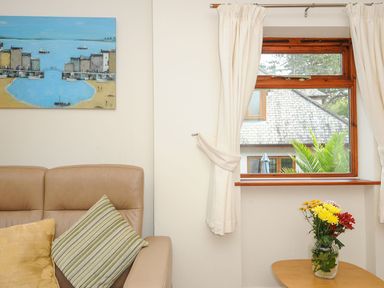 A beige sofa with two cushions next to a window with cream curtains and a wooden table with a vase of flowers at Little Trevallan in Looe