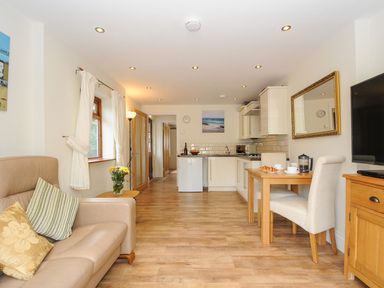 A living area with a beige sofa and pillows a wooden dining table with chairs a kitchen with white cabinets and appliances and a television on a wooden stand at Little Trevallan in Looe