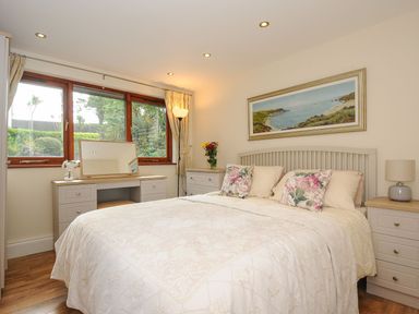 A bedroom with a bed, floral pillows, dresser, mirror, lamps, and a window at Little Trevallan in Looe