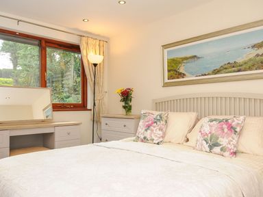 A bedroom with a bed and floral pillows a desk with a mirror a floor lamp and a landscape painting at Little Trevallan in Looe