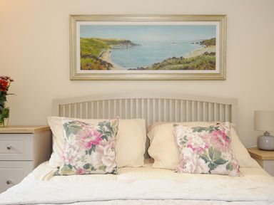A bedroom with a bed with floral pillows a painting above and bedside tables with a lamp and flowers at Little Trevallan in Looe