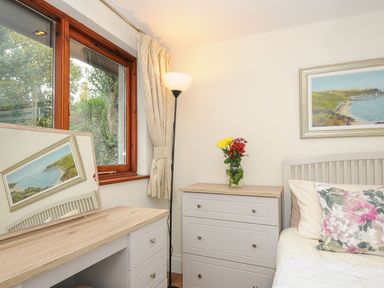 A bedroom corner with window curtains floor lamp dresser with flowers and bed with floral pillow at Little Trevallan in Looe