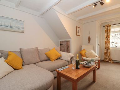 A living room with a grey sofa and yellow cushions a wooden coffee table with a bottle and glasses and a white armchair with cushions at Shell Seeker Cottage in Sewerby