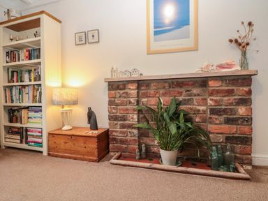 A brick fireplace with a potted plant and glass bottles next to a wooden chest with a lamp and a bookshelf in a room at Shell Seeker Cottage in Sewerby