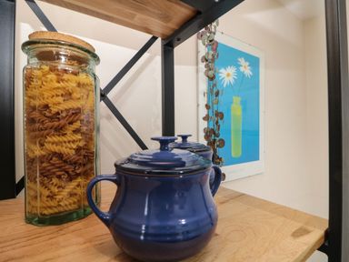A kitchen shelf with a jar of pasta and two blue pots next to a wall with a framed flower print at Shell Seeker Cottage in Sewerby