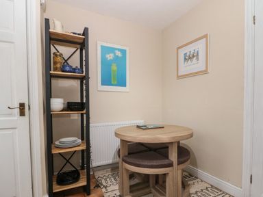A small dining area with a round wooden table and four stools a shelving unit with dishes and jars and framed pictures on beige walls at Shell Seeker Cottage in Sewerby