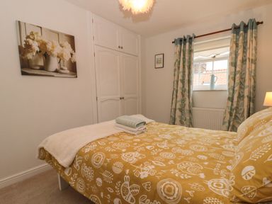 A bedroom with a bed covered in yellow patterned bedding two folded towels on the bed a window with patterned curtains and white built in wardrobes at Shell Seeker Cottage in Sewerby