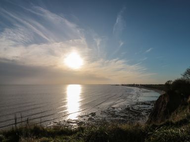 A coastal view with a sun reflecting on the water and a shoreline with rocks and vegetation at Shell Seeker Cottage in Sewerby