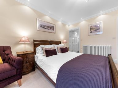 A bedroom with a bed with white and purple bedding a wooden nightstand with a lamp a purple armchair and framed pictures on the wall at Gate House in Coniston