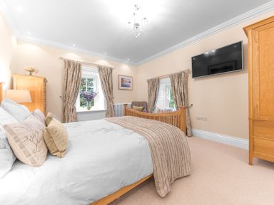 A bedroom with a wooden bed with pillows and a blanket a wooden wardrobe a wall-mounted TV two windows with curtains and a chair at Gate House in Coniston
