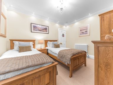 A bedroom with two single wooden beds a wooden nightstand with a lamp a wooden dresser framed pictures on the walls and a white door at Gate House in Coniston