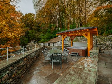 An outdoor stone patio with metal dining table and chairs next to a covered hot tub surrounded by trees and stone walls at Gate House in Coniston